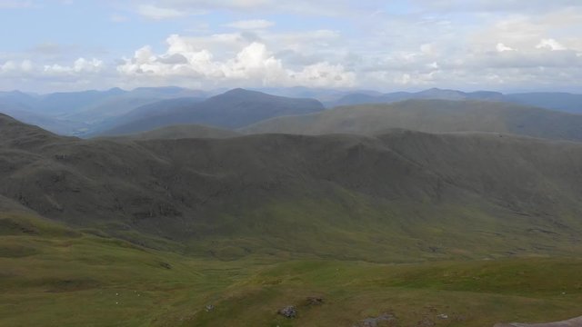 An aerial backward footage of a grassy mountain rocky summit with small pond and mountain range in the background under a cloudy sky