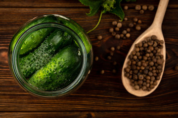 Fresh cucumbers in glass jar, garlic and black pepper on dark wooden background.