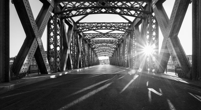 Black And White Asphalt Road Under The Steel Construction Of A Bridge In The City On A Sunny Day. Evening Urban Scene With The Sunbeam In The Tunnel. City Life, Transport And Traffic Concept.	