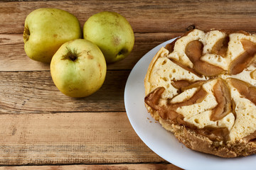 Baked apple pie on a white plate next to three green apples on a wooden background