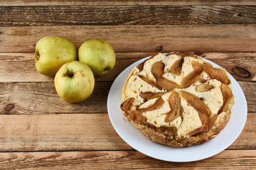 Naklejka na ściany i meble Baked apple pie on a white plate next to three green apples on a wooden background