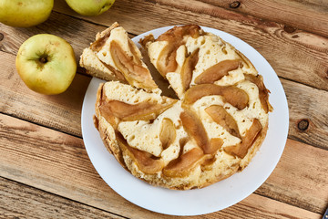 Sliced piece of baked apple pie on a white plate next to green apples on a wooden background