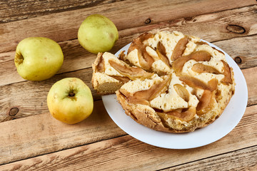 Naklejka na ściany i meble Sliced piece of baked apple pie on a white plate next to green apples on a wooden background