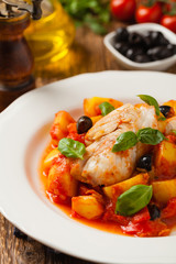 Traditionally prepared cod in Spanish. Served in tomato sauce with boiled potatoes, olivki and basil. Front view.