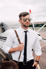 confident pilot in sunglasses with bag standing with hand in pocket near plane