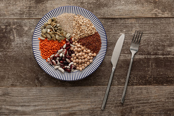 Obraz na płótnie Canvas top view of fork and knife near striped ceramic plate with raw assorted beans, cereals and seeds on dark wooden surface with copy space