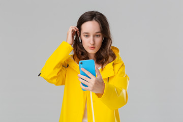 Pretty brunette woman in a yellow raincoat is making selfie with the smartphone isolated over grey background. Model wears wireless earbuds. Mobile photography.