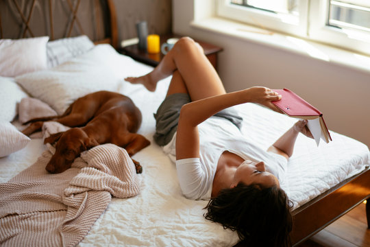 Young woman with dog  lying on bed while reading book