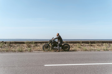 Fototapeta na wymiar Brutal man in leather jacket and sigar sitting on bike near ocean