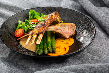 Roasted rack of lamb served with asparagus, yellow tomatoes and lamb sauce. Loin lamb Served in black stone plate with fork and knife. Gray background. Tuna salad. Top view from above horizontal.