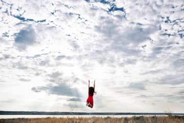 Back view of a jumping long haired girl in a red dress on a cloudy day near the lake