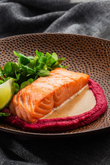 Fried fish salmon fillet with pink humus garnish. One piece of baked salmon grilled lime, tomatoes and salade on a brown plate . Gray background. Horizontal photo.