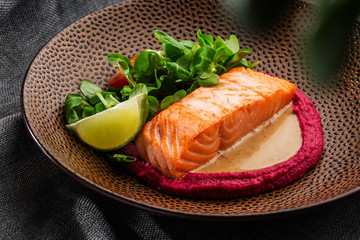Fried fish salmon fillet with pink humus garnish. One piece of baked salmon grilled lime, tomatoes and salade on a brown plate . Gray background. Horizontal photo.