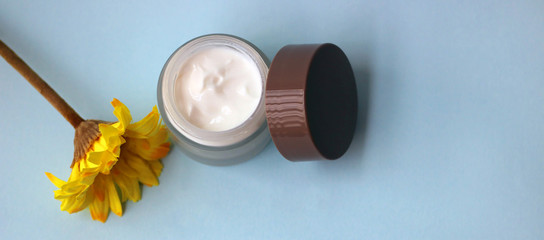 Jar of moisturizer for the face next to a yellow flower on a light blue background.