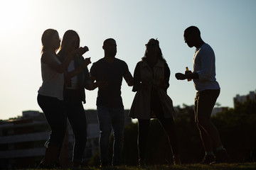 Friends enjoying outdoor party at sunset. Silhouettes of multiethnic men and women drinking beer and dancing on grass. Party at sunset concept