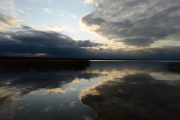 Clouds in the blue sky reflect on the calm clear water of the lake on sunset