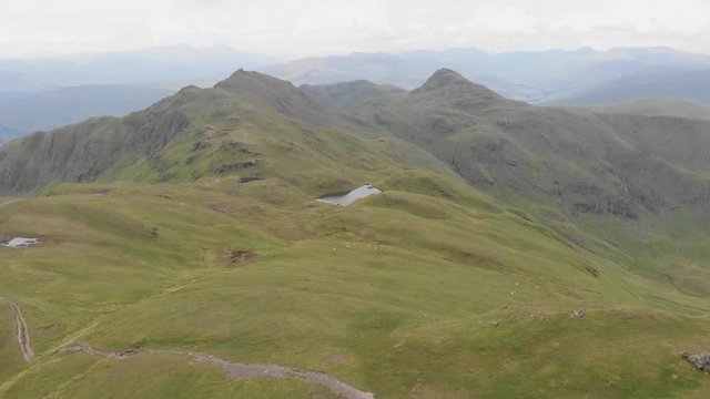 An aerial forward footage of a grassy mountain plateau rocky summit with small pond and mountain range in the background under a cloudy sky