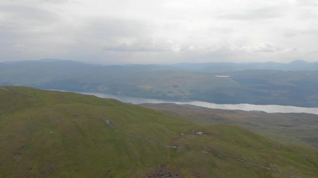 An aerial sideway right to left footage of a grassy mountain plateau summit with a huge lake and mountain range in the background under a cloudy sky	