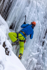 climber in a blue jacket, yellow pants and a red helmet climbs the frozen waterfall. ice climbing the waterfall. rear view. vertical view