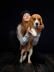 A red-haired girl of ten years old plays with a beagle puppy sitting on a dark wooden floor opposite a black background.