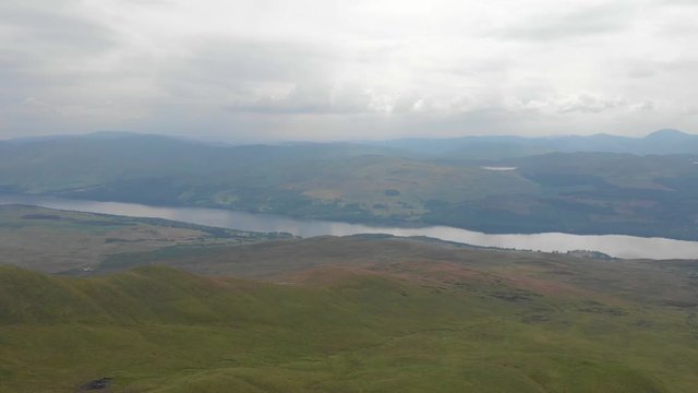 An aerial backward footage of a grassy mountain plateau with a huge lake and mountain range in the background under a cloudy sky	