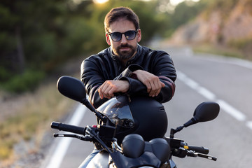 Handsome bearded biker looking at camera while sitting on his motorbike and holding helmet.