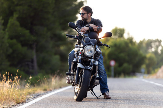 Handsome bearded biker looking to sideways while sitting on his motorbike and holding helmet.