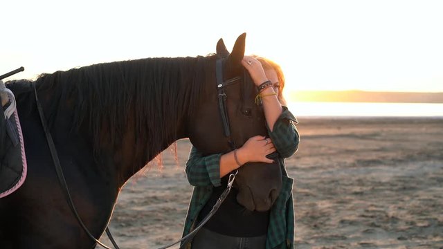 Blonde woman stroking and hugging horse. Beautiful lady with black stallion enjoying sunset nature. Love and friendship concept. Slow motion.