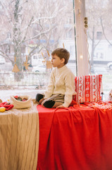 Cute little boy in a warm white knitted sweater sits at home by the window decorated for Christmas...