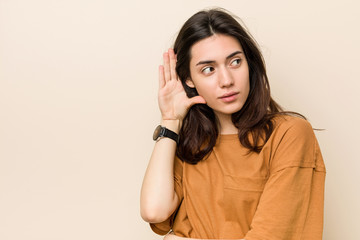 Young brunette woman against a beige background trying to listening a gossip.