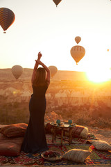 a girl in a long dark dress is standing on the carpet in front of sunrise and air balloons in the sky