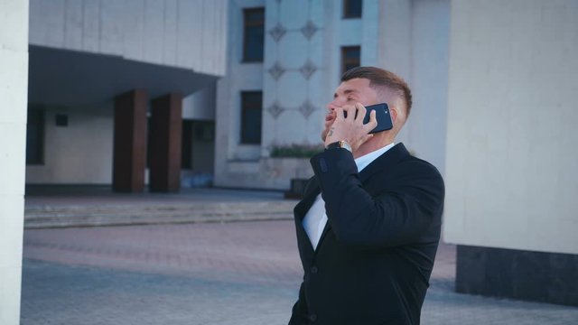 Young successful businessman in suit, talking on the phone, success in deal, smiling, in the background of the office building. Good luck in business, successful deal, good news.