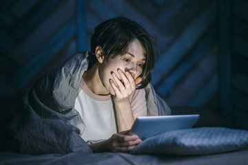 Portrait of a young woman watching a horror movie at home. She covers her mouth with her hand in...