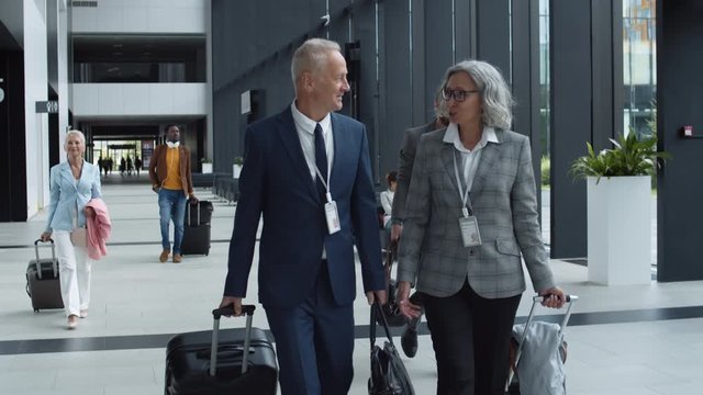 Medium shot of old good-looking Caucasian businessman and businesswoman with suitcases going together in airport and communicating