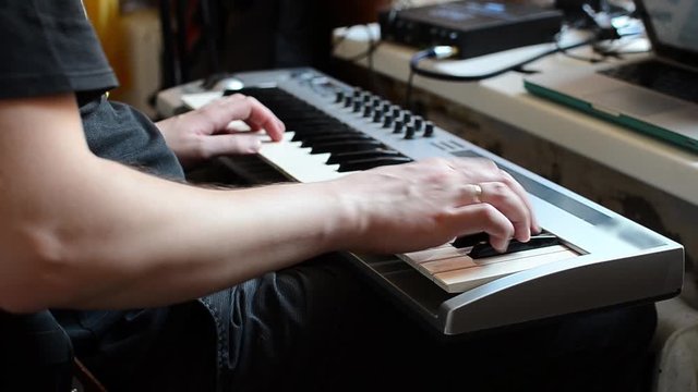 A white man in a black t-shirt plays his favorite tune on a synthesizer. The musician plays the piano