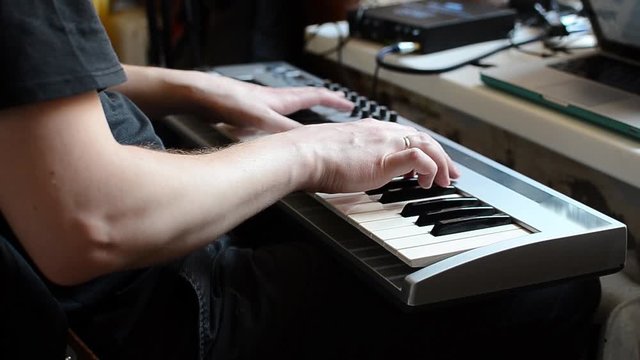 A white man in a black t-shirt plays his favorite tune on a synthesizer. The musician plays the piano