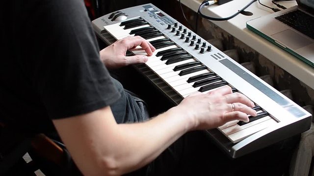 A white man in a black t-shirt plays his favorite tune on a synthesizer. The musician plays the piano