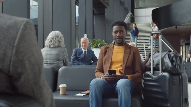 Tracking left of African man wearing casual clothes sitting in airport lounge with smartphone in his hands, looking at camera and smiling