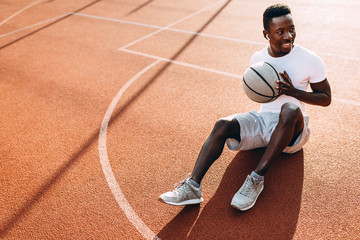 Sporty African American resting after a workout sitting on the court with a basketball, basketball player