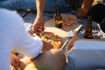 Hand of man taking slice of pizza from box. People gathering around plaid with food and drink, sitting on grass, drinking beer. Eating outdoors concept