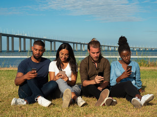 Focused young men and women sitting in line on grass and using smart phones. Diverse team of friends obsessed with gadgets. Social media addiction concept