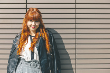 Cute young girl in a black leather jacket with curly red hair and a fringe stands near the modern wall on a sunny day and looks at the camera