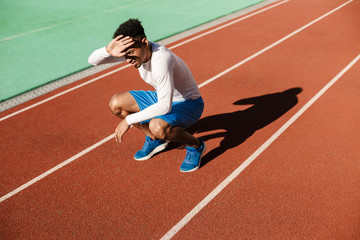 Young tired African American sportsman sit on one's haunches covering face from sun after run at...
