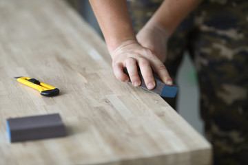 Fototapeta na wymiar Focus on male hands holding special tool and grinding surface of desk. Manufacture of stylish wooden furniture. Carpentry workshop concept. Blurred background