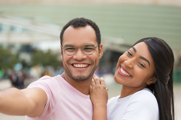 Cheerful young couple posing for selfie on street. Happy multiracial friends smiling for self portrait. African American man with outstretched arm. Concept of self portrait