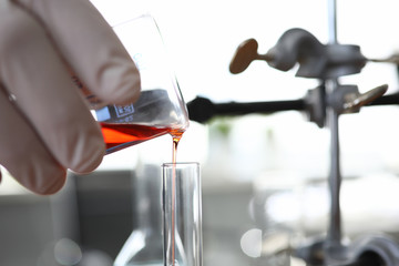 Focus on male hand in gloves holding glass flask. Pharmacist transfusing red fluid in bulb. Medical research of new drugs concept. Laboratory interior on blurred background