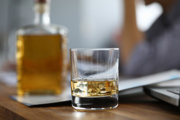 Focus on glassful with alcoholic beverage with ice-cube on wooden brown table. Relax after tough day. Scotch or whisky gourmet alcohol. Blurred background