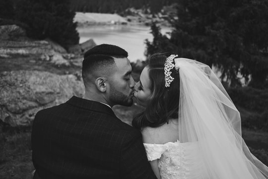 Black and white wedding photo of loving Kazakh Asian couple in nature, beautiful bride in dress and veil and groom in suit hug and kiss