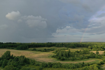 Top view of meadow with road and green plants. Cloudy sky and beautiful rainbow above trees and bushes. Picturesque landscape scene of rain in summer time