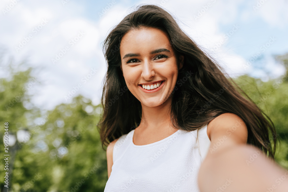 Canvas Prints Beautiful young brunette smiling woman taking self portrait on nature and sky background in the park. Pretty female making selfie on her device outdoors. People, lifestyle and travel